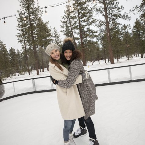 Two women dressed in warm winter gear hugging while ice skating outdoors in the snow.